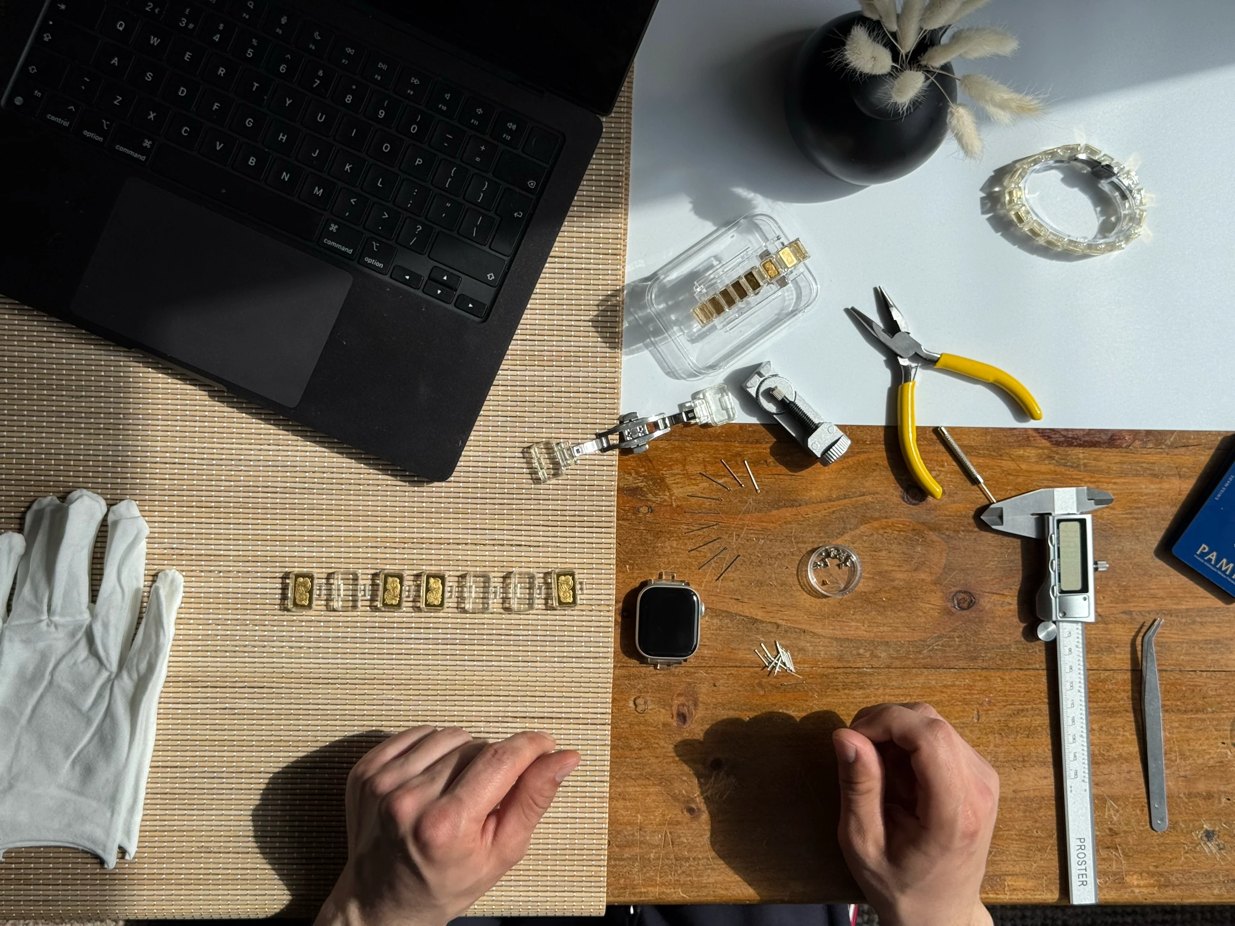 A workbench seen from above: a row of capsules, a laptop, pliers and callipers, with two hands at work