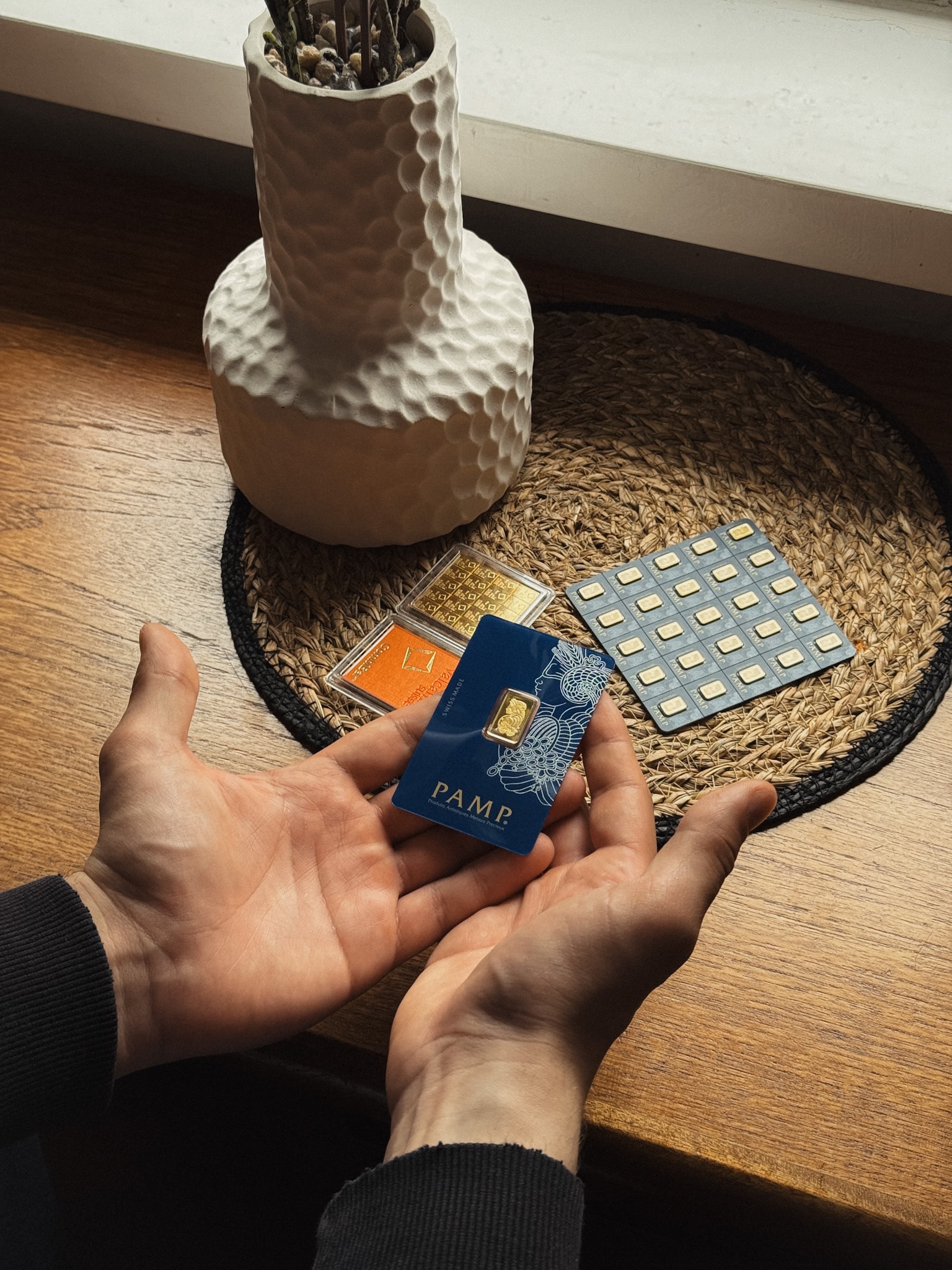 Hands holding gold bars in PAMP protective cards on a wooden surface